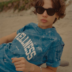 Person wearing a blue 'Wellness' shirt sitting on a beach.
