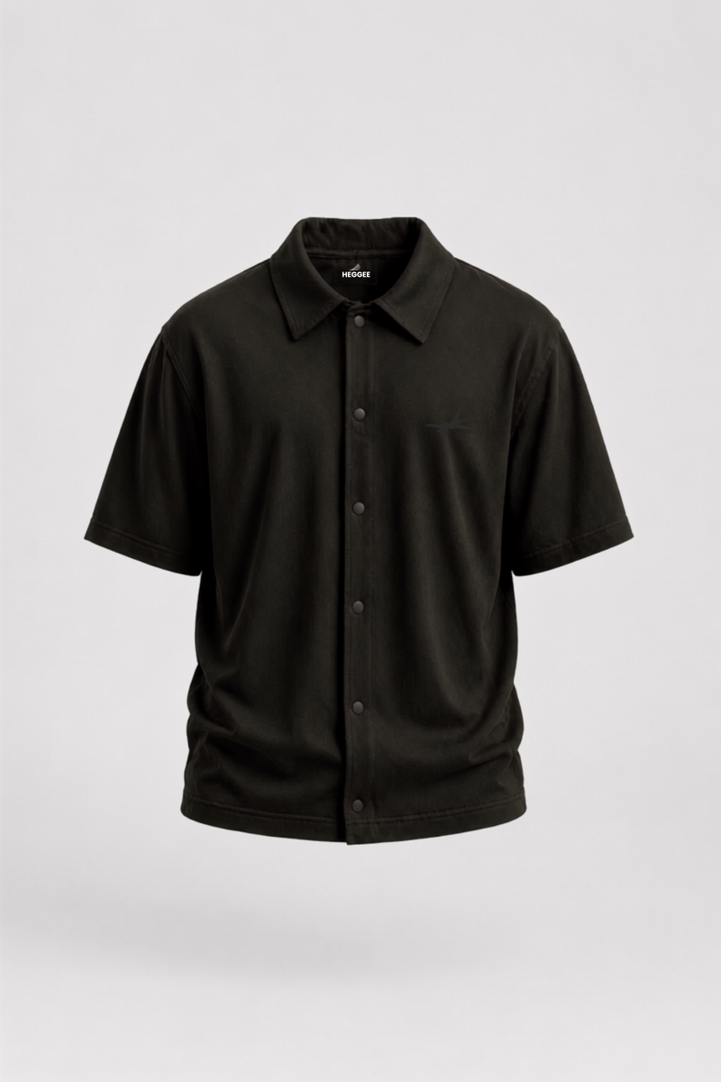 A black short-sleeve button-up camp shirt with a classic collar and a single chest pocket, displayed on a neutral light gray background.