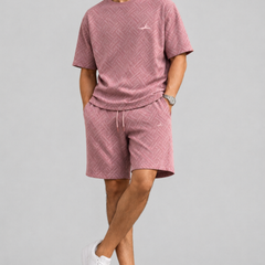 A man modeling a dusty pink textured two-piece lounge set consisting of a short-sleeve crewneck t-shirt and matching drawstring shorts, paired with white sneakers, against a solid light gray background.