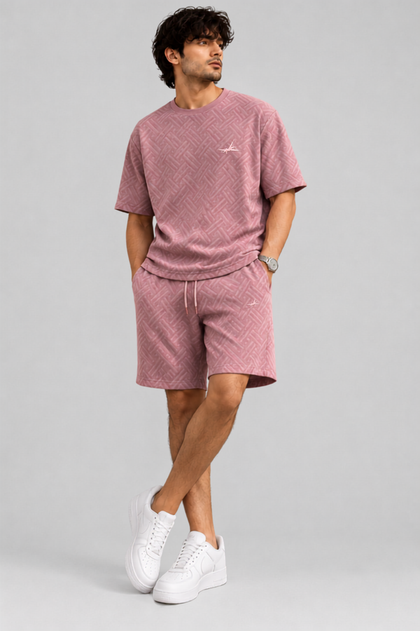 A man modeling a dusty pink textured two-piece lounge set consisting of a short-sleeve crewneck t-shirt and matching drawstring shorts, paired with white sneakers, against a solid light gray background.