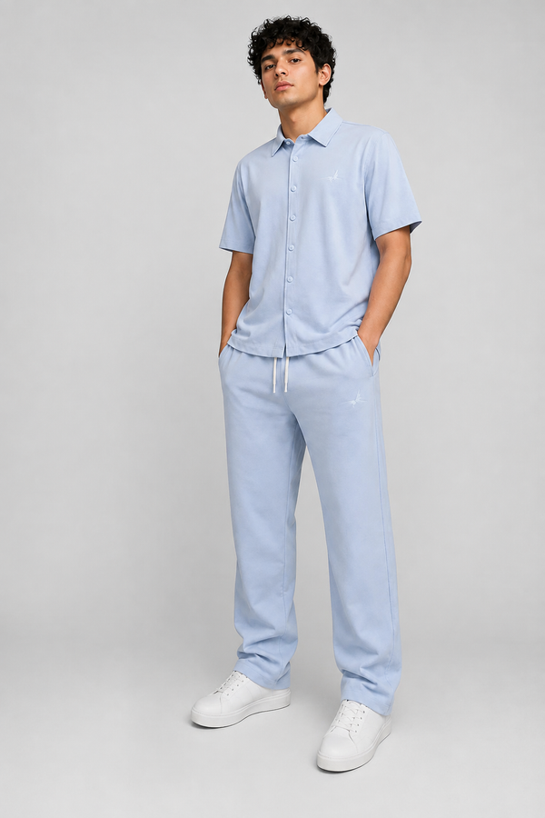 A man modeling a light blue short-sleeve button-up shirt and matching light blue jogger pants with white sneakers, standing against a neutral light gray studio background.