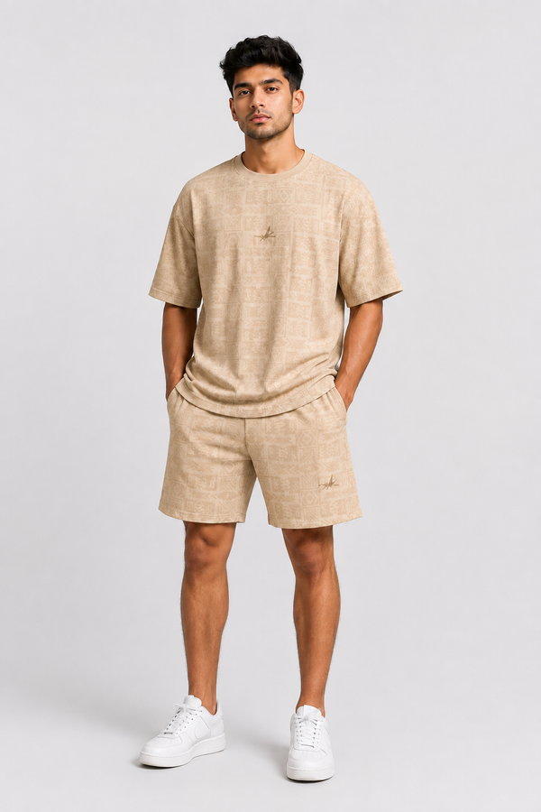 A man modeling a matching beige textured two-piece lounge set consisting of a short-sleeve t-shirt and shorts, paired with white sneakers, against a light gray studio background.
