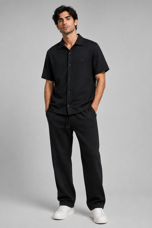 A man modelling a black short-sleeve button-up shirt and matching black drawstring sweatpants against a solid grey studio background.