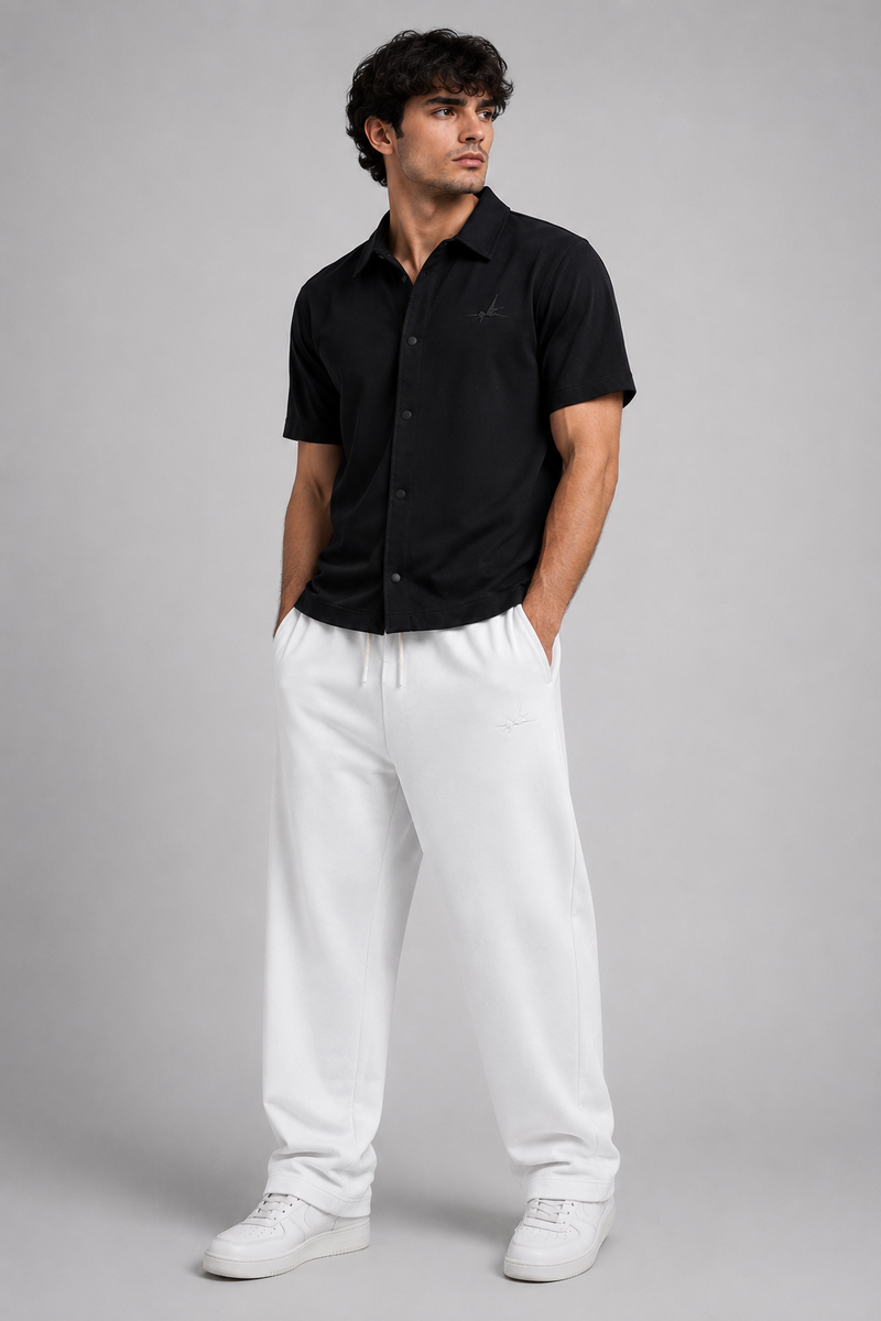 A man modelling a black short-sleeve button-up shirt and white sweatpants against a solid gray studio background.
