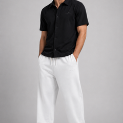 A man modelling a black short-sleeve button-up shirt and white sweatpants against a solid gray studio background.
