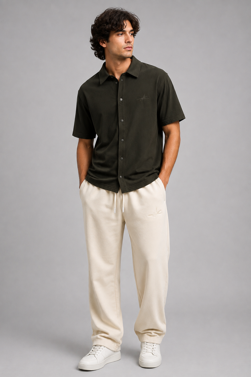 A man modelling a dark olive green short-sleeve button-up shirt and cream-colored sweatpants against a solid gray studio background.