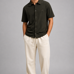 A man modelling a dark olive green short-sleeve button-up shirt and cream-colored sweatpants against a solid gray studio background.