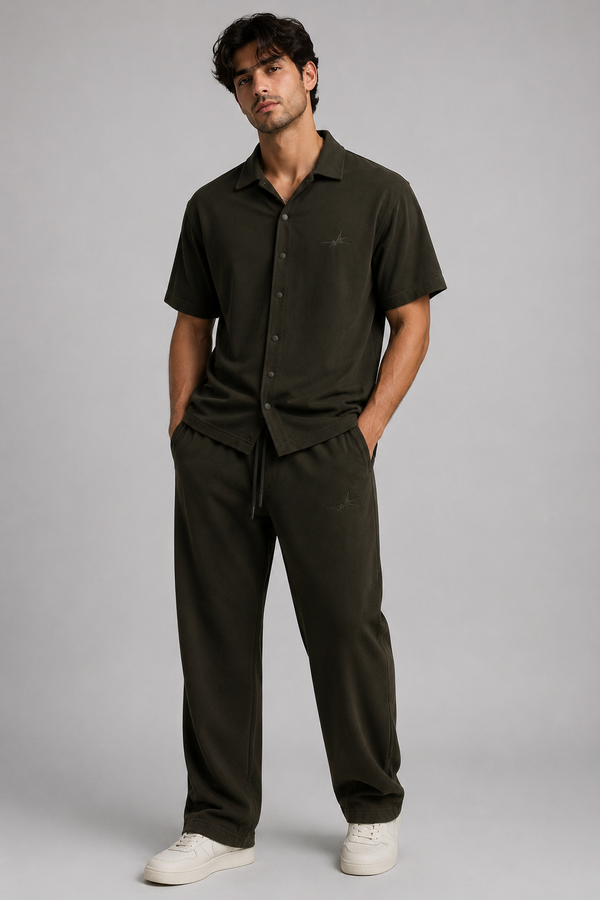 A man modelling a dark olive green short-sleeve button-up shirt and matching drawstring sweatpants against a solid gray studio background.