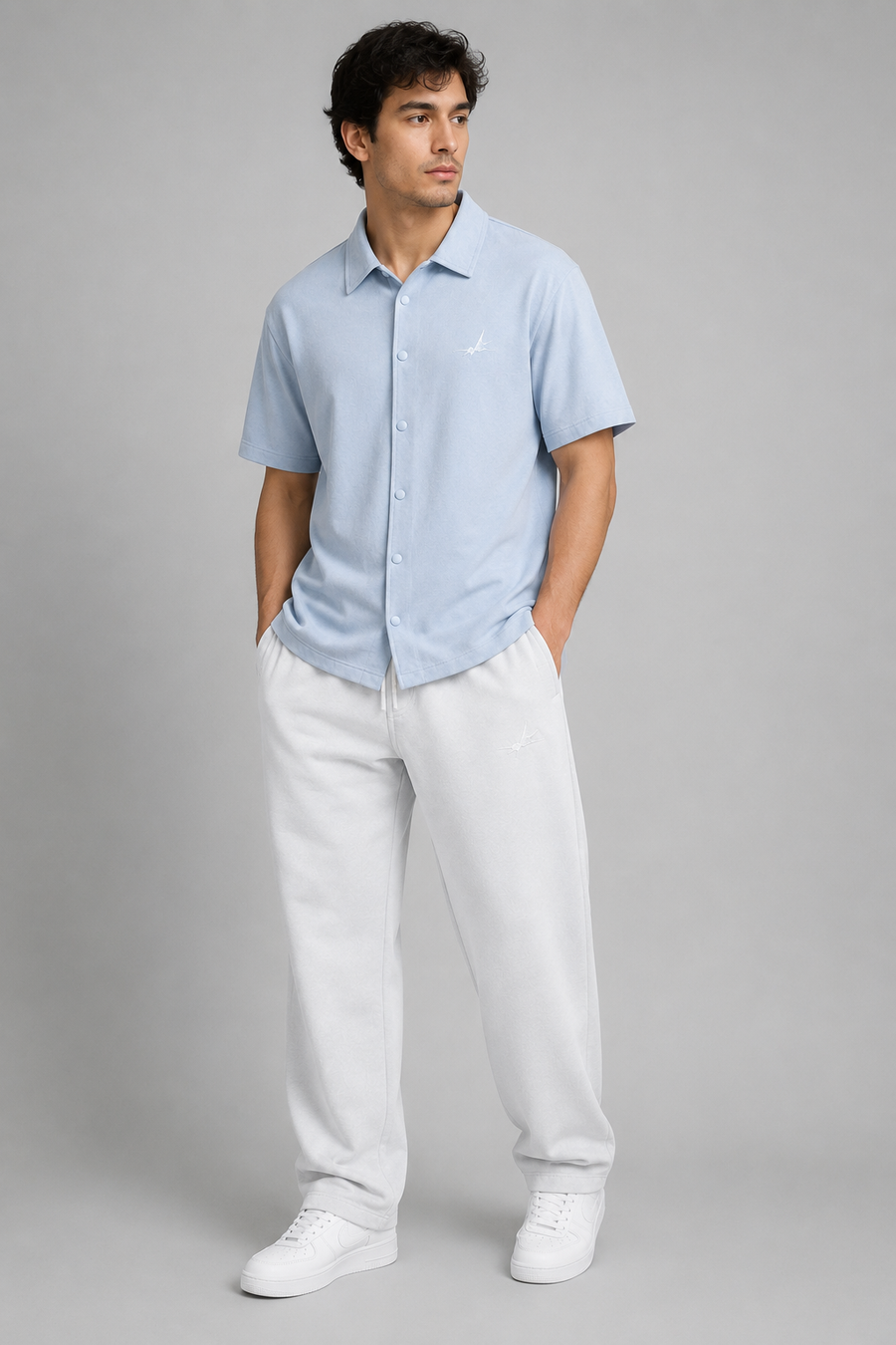 A man modelling a light blue short-sleeve button-up shirt and white sweatpants against a solid gray studio background.