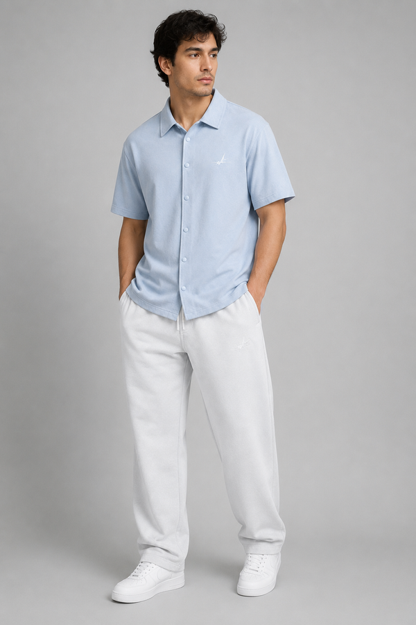 A man modelling a light blue short-sleeve button-up shirt and white sweatpants against a solid gray studio background.
