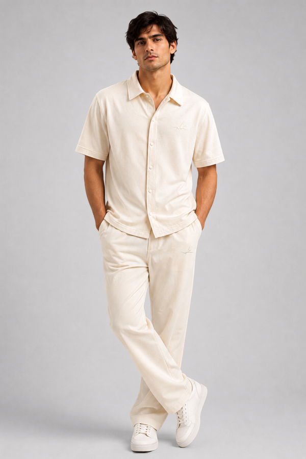 A man modelling a matching cream-colored short-sleeve button-up shirt and trousers set, paired with white sneakers, against a solid light gray studio background.