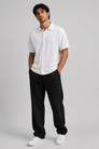 A man modelling a white short-sleeve button-up shirt and black sweatpants against a light grey studio background.