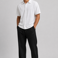 A man modelling a white short-sleeve button-up shirt and black sweatpants against a light grey studio background.