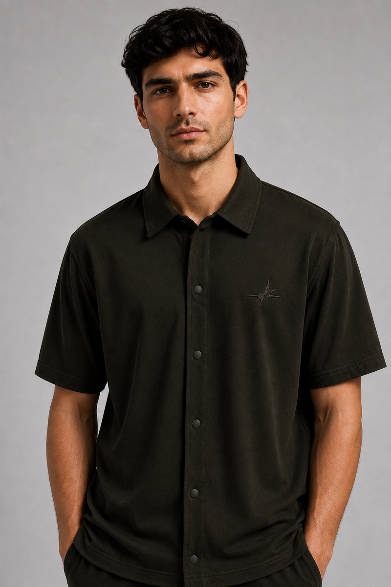 A man wearing a dark olive green short-sleeve button-up shirt with a small embroidered logo on the left chest, photographed against a neutral gray studio background.