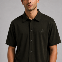 A man wearing a dark olive green short-sleeve button-up shirt with a small embroidered logo on the left chest, photographed against a neutral gray studio background.