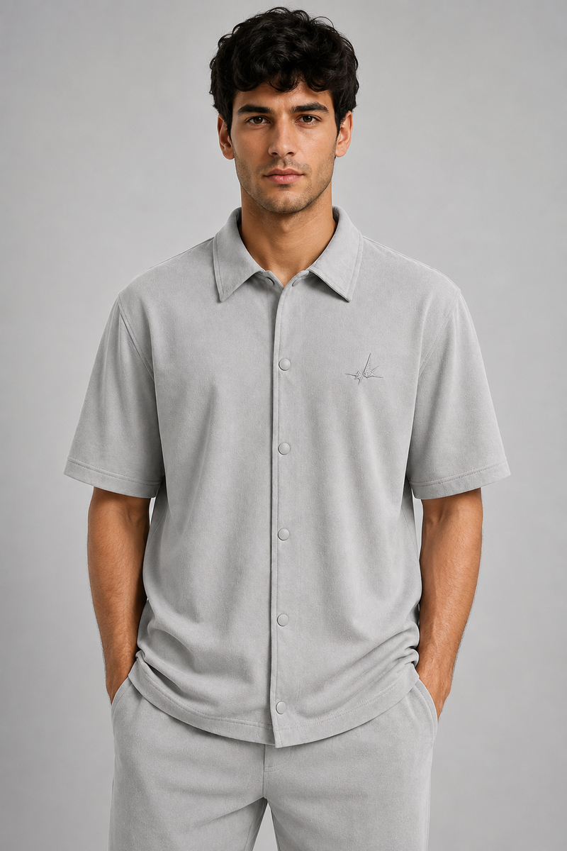 A man wearing a light gray short-sleeve button-up polo shirt with a small embroidered logo on the chest, paired with matching gray trousers, against a solid gray studio background.