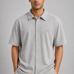 A man wearing a light gray short-sleeve button-up polo shirt with a small embroidered logo on the chest, paired with matching gray trousers, against a solid gray studio background.