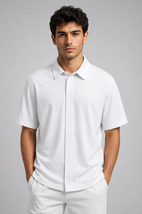 A man wearing a white short-sleeve button-up shirt and matching white trousers, standing against a solid gray studio background.