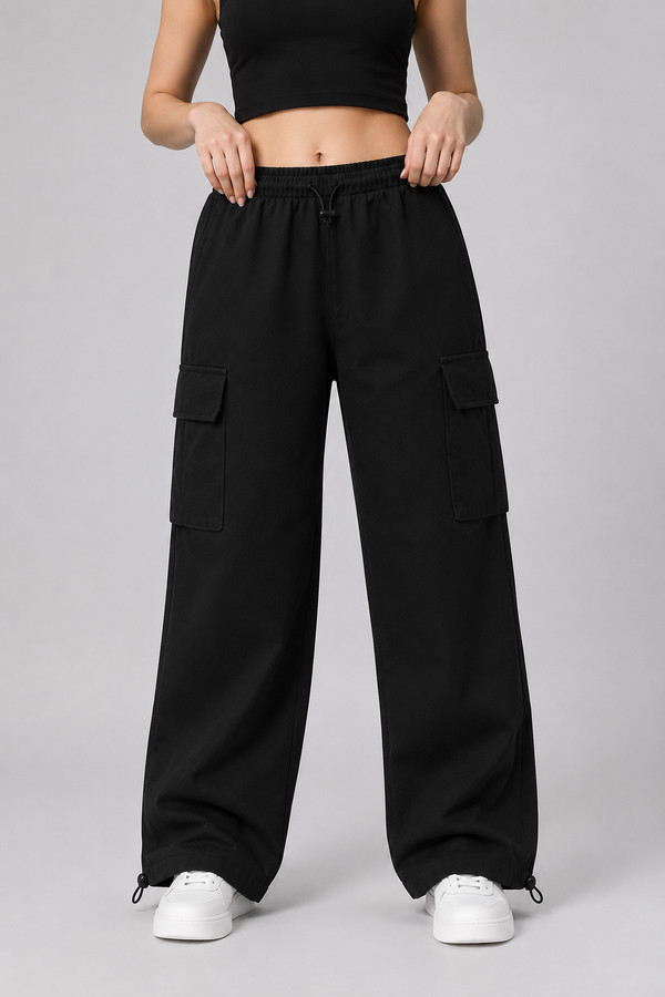 A model wearing black wide-leg cargo pants with side flap pockets, paired with a black crop top and white sneakers, shown from the waist down against a neutral gray studio background.
