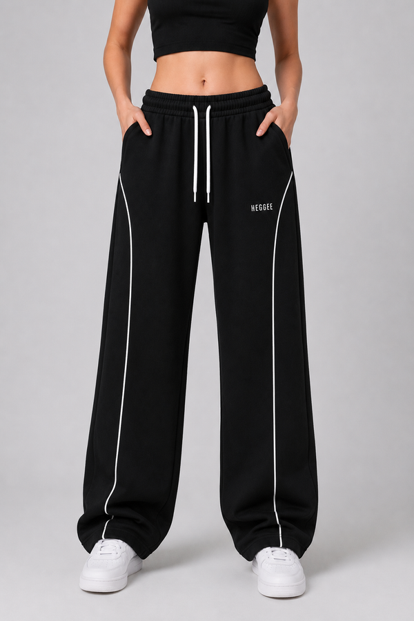 A model wearing black wide-leg sweatpants with white side piping and a white drawstring, paired with white sneakers against a light gray studio background.