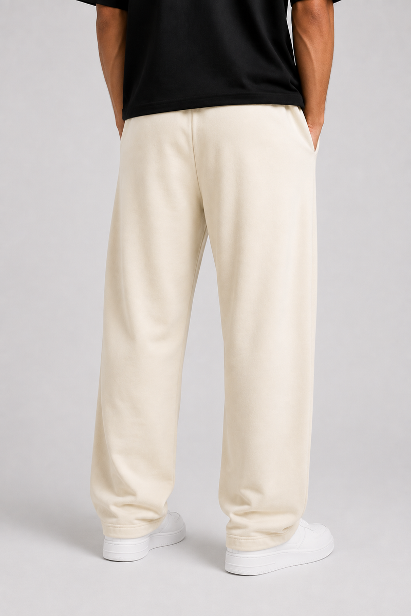 A person shown from the back wearing loose, straight-leg cream or off-white sweatpants, a black top, and white sneakers against a light gray studio background.