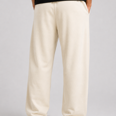 A person shown from the back wearing loose, straight-leg cream or off-white sweatpants, a black top, and white sneakers against a light gray studio background.