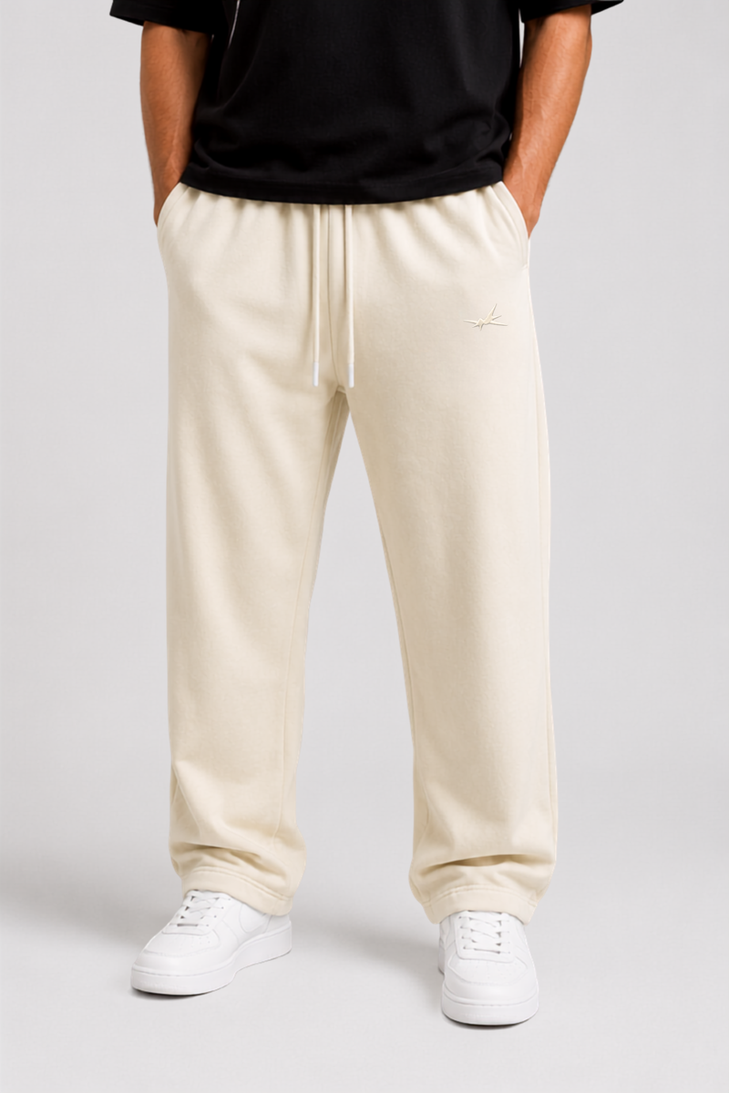 A person wearing beige drawstring sweatpants with a small embroidered logo on the left thigh, paired with a black top and white sneakers, shown from the waist down against a light gray studio background.