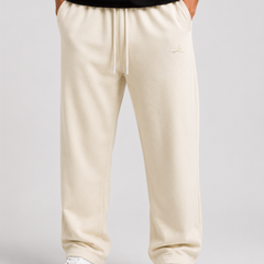 A person wearing beige drawstring sweatpants with a small embroidered logo on the left thigh, paired with a black top and white sneakers, shown from the waist down against a light gray studio background.