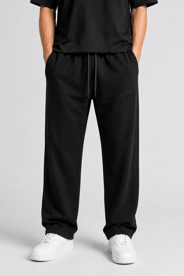 A person wearing black drawstring sweatpants with a small tonal embroidered logo on the left thigh, paired with a black top and white sneakers, photographed against a light gray studio background.