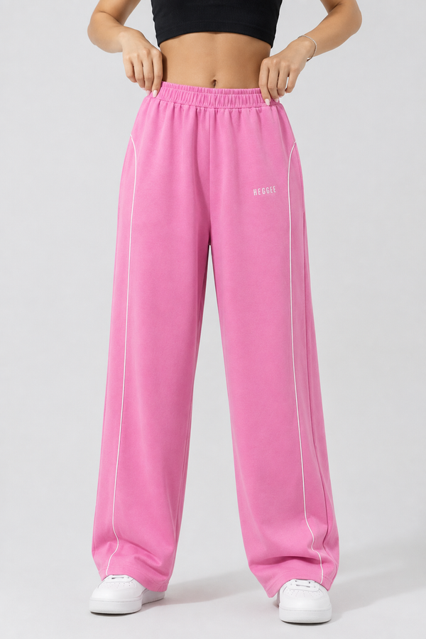 A person wearing bright pink wide-leg sweatpants with white piping and a small white logo on the upper left thigh, paired with white sneakers against a light gray studio background.