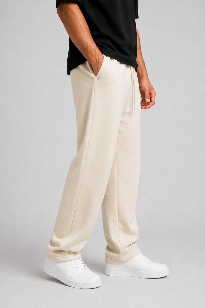 A person wearing cream-colored sweatpants, a black t-shirt, and white sneakers, standing against a light gray studio background.