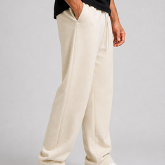A person wearing cream-colored sweatpants, a black t-shirt, and white sneakers, standing against a light gray studio background.