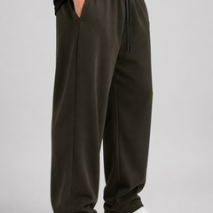 A person wearing dark olive green drawstring sweatpants with a black top and white sneakers, standing against a light gray studio background.