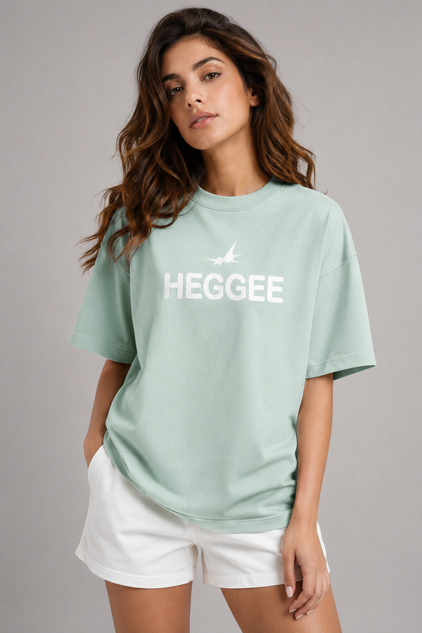 A woman modelling a light mint green graphic t-shirt with the word 'HEGGEE' and a small airplane logo printed in white across the chest, paired with white shorts against a solid gray studio background.