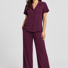 A woman modelling a two-piece pajama set in deep plum purple, featuring a short-sleeve button-front top and matching wide-leg pants, photographed against a light gray studio background.