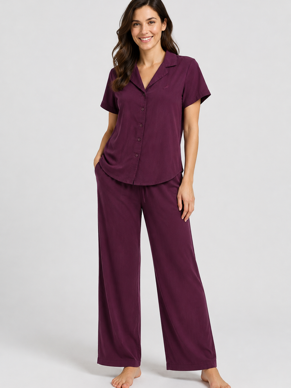 A woman modelling a two-piece pajama set in deep plum purple, featuring a short-sleeve button-front top and matching wide-leg pants, photographed against a light gray studio background.
