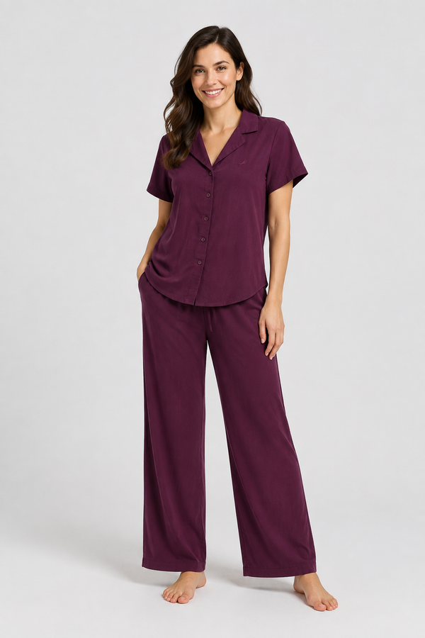 A woman modelling a two-piece pajama set in deep plum purple, featuring a short-sleeve button-front top and matching wide-leg pants, photographed against a light gray studio background.