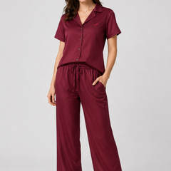 A woman modelling a two-piece sleepwear set consisting of a short-sleeve button-front top and matching drawstring pants in a deep burgundy color, shot against a solid light gray studio background.