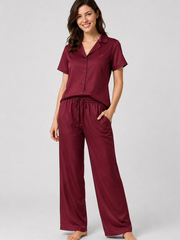 A woman modelling a two-piece sleepwear set consisting of a short-sleeve button-front top and matching drawstring pants in a deep burgundy color, shot against a solid light gray studio background.