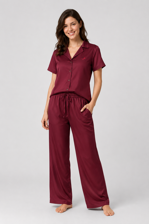 A woman modelling a two-piece sleepwear set consisting of a short-sleeve button-front top and matching drawstring pants in a deep burgundy color, shot against a solid light gray studio background.