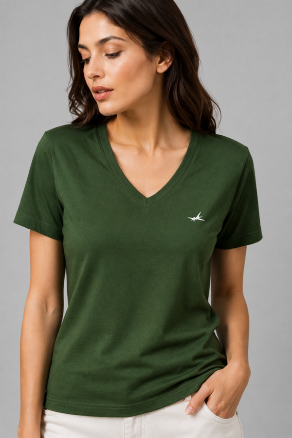 A woman wearing a dark green V-neck short-sleeve t-shirt with a small white bird logo on the left chest, paired with white pants, set against a solid gray studio background.