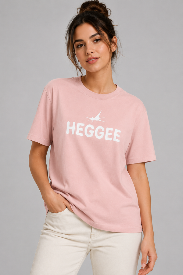 A woman wearing a light pink crewneck t-shirt with a white graphic print that reads 'HEGGEE' and a small airplane silhouette, paired with off-white pants, against a solid gray studio background.