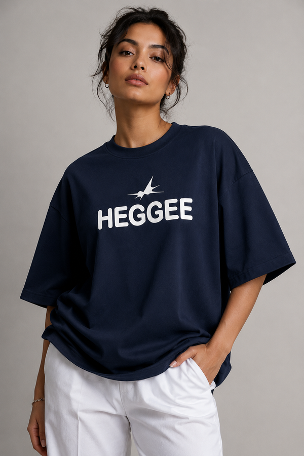 A woman wearing an oversized navy blue t-shirt with a white 'HEGGEE' text and bird logo on the chest, paired with white pants, against a neutral gray background.